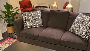 A living room with a dark brown sofa, two patterned throw pillows, a glass coffee table, and a remote control on it. There are two lamps with black shades, a potted plant, and a red throw blanket on the sofa. - Downtown!  Great neighborhood!  Walk everywhere downtown!   (Anchorage)