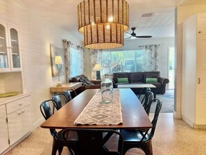 A dining area with a wooden table, black chairs, a woven pendant light, and a view of a living room with a sofa and curtains. - Charming Sarasota Home • Near Beaches & Downtown, 3bed, 2 bath. (Sarasota)
