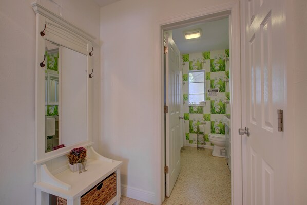 A hallway leading to a bathroom with a white door, a mirror, and a small white console table with a vase. - Charming Sarasota Home • Near Beaches & Downtown, 3bed, 2 bath. (Sarasota)