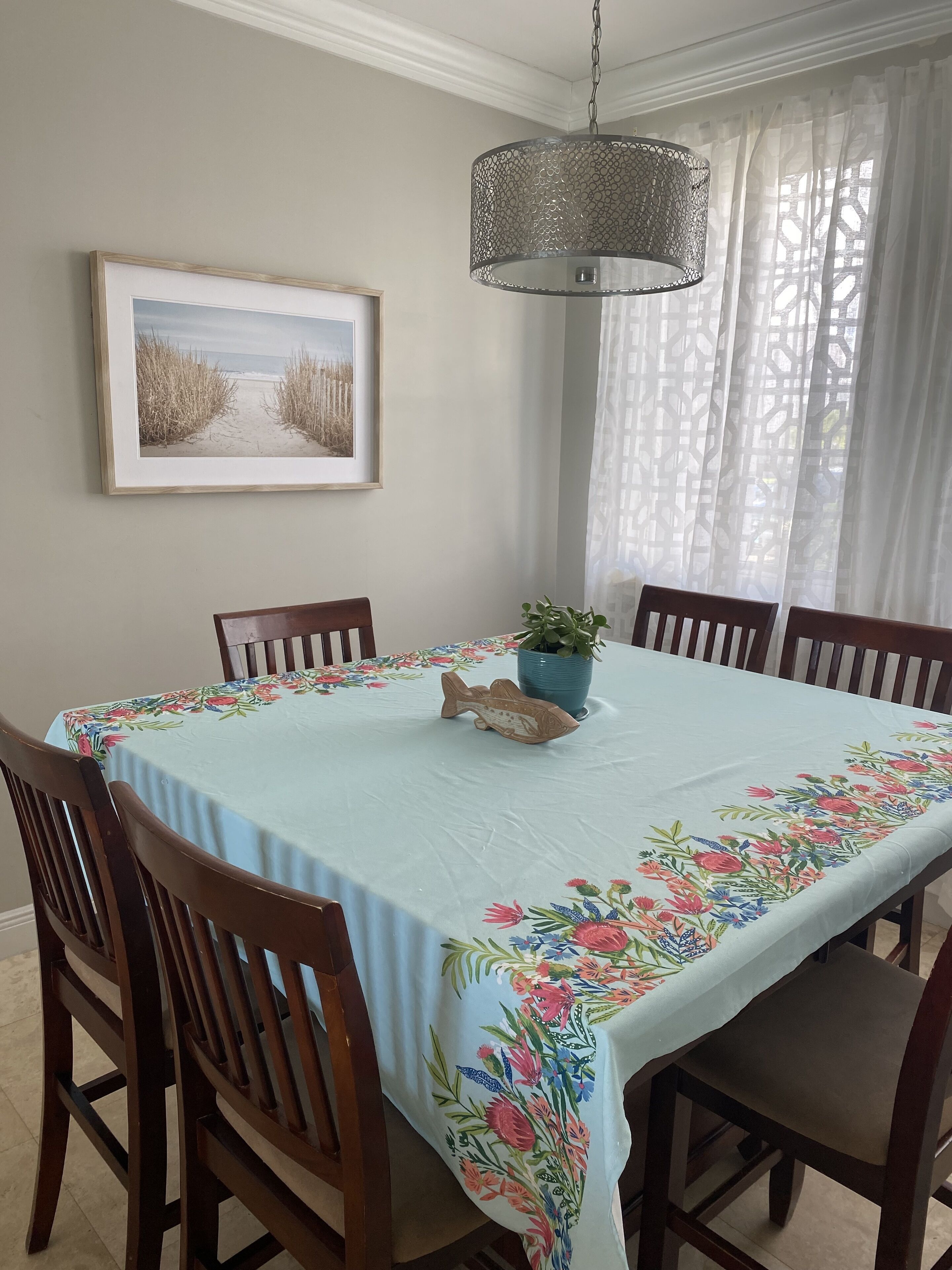 A dining area with a table covered by a floral tablecloth, surrounded by wooden chairs, a hanging light fixture, and a framed picture on the wall.