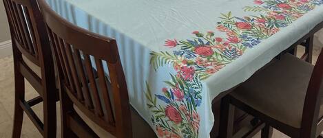 A dining area with a table covered by a floral tablecloth, surrounded by wooden chairs, a hanging light fixture, and a framed picture on the wall.