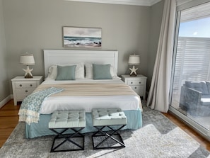 A bedroom with a large bed, two nightstands, star-shaped lamps, a beach-themed painting, and a view of the outdoors.