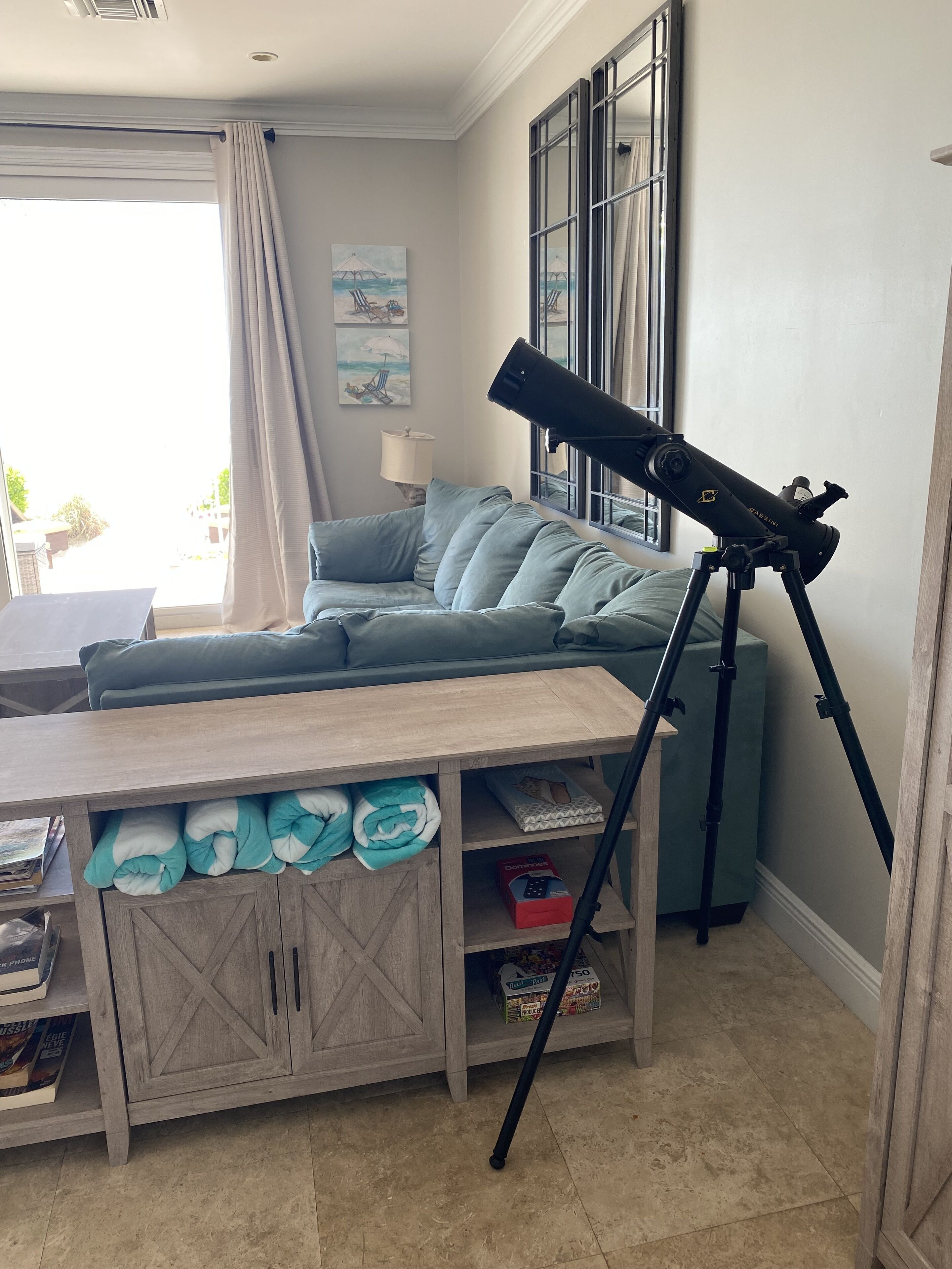 A living room with a telescope on a tripod, a sofa, a wooden table with cushions, and a window with curtains.