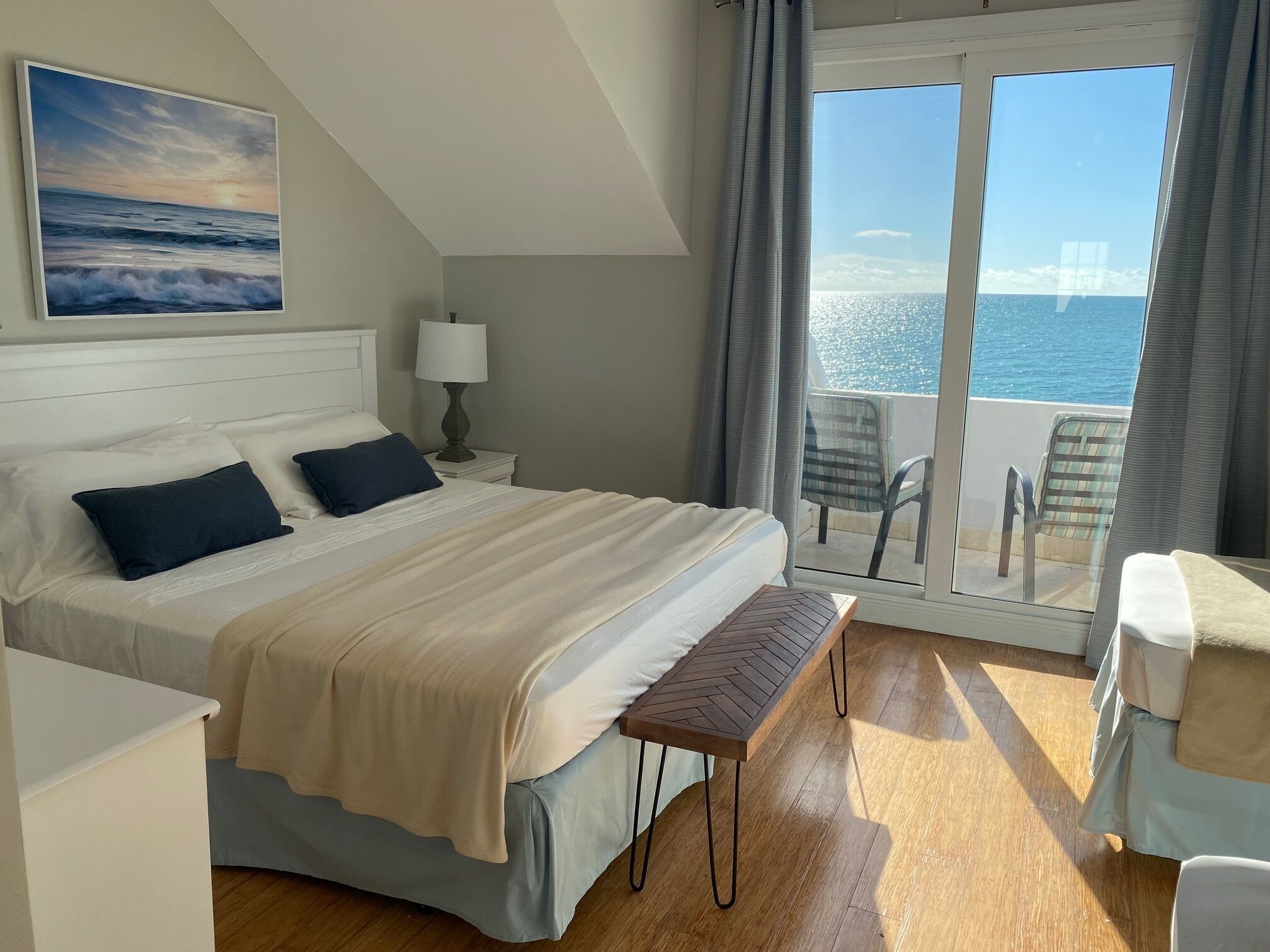 A hotel room with a large bed, a bench, a bedside table with a lamp, a view of the sea through a sliding door, and a painting on the wall.