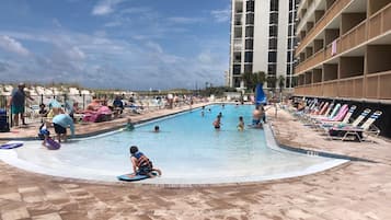 A swimming pool area with multiple pools, lounge chairs, and a high-rise building in the background.