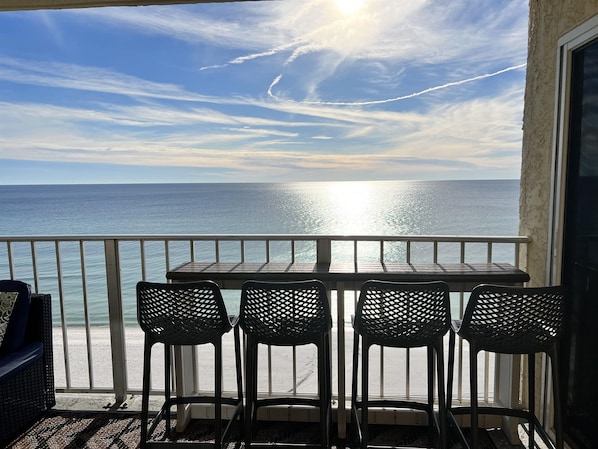 A balcony with a table and chairs overlooking the sea, with a clear sky and visible contrails.
