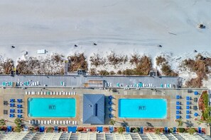 An aerial view of a resort with two large swimming pools, a parking area, and a beach.