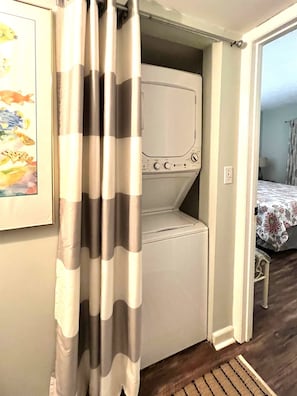 A compact laundry room with a washing machine and dryer, a striped curtain, and a bed with a floral bedspread in the adjacent room.