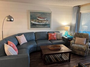 Ein Wohnzimmer mit einer gebogenen Couch, einem Couchtisch, einem Sessel, einer Lampe und einem Gemälde von Booten an der Wand.