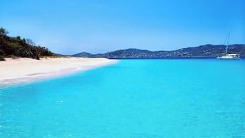 A beach with clear turquoise water, a sandy shore, and a sailboat in the distance.