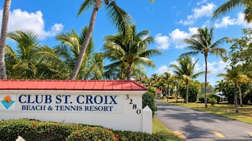 A sign for Club St. Croix Beach & Tennis Resort with a red roof and white walls, surrounded by palm trees and a clear sky.
