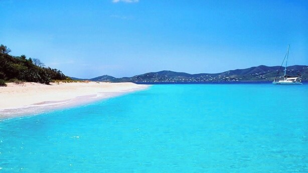 Ein Strand mit klarem türkisfarbenem Wasser, einem Sandufer und einem Segelboot in der Ferne.