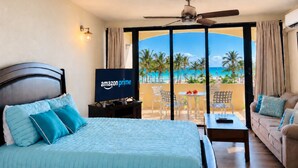 Hotelkamer met een bed, een televisietoestel waarop het Amazon Prime logo te zien is, een balkon met uitzicht op het strand en een plafondventilator.