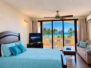 Habitación de hotel con cama, televisor mostrando el logotipo de Amazon Prime, balcón con vista a la playa y ventilador de techo.