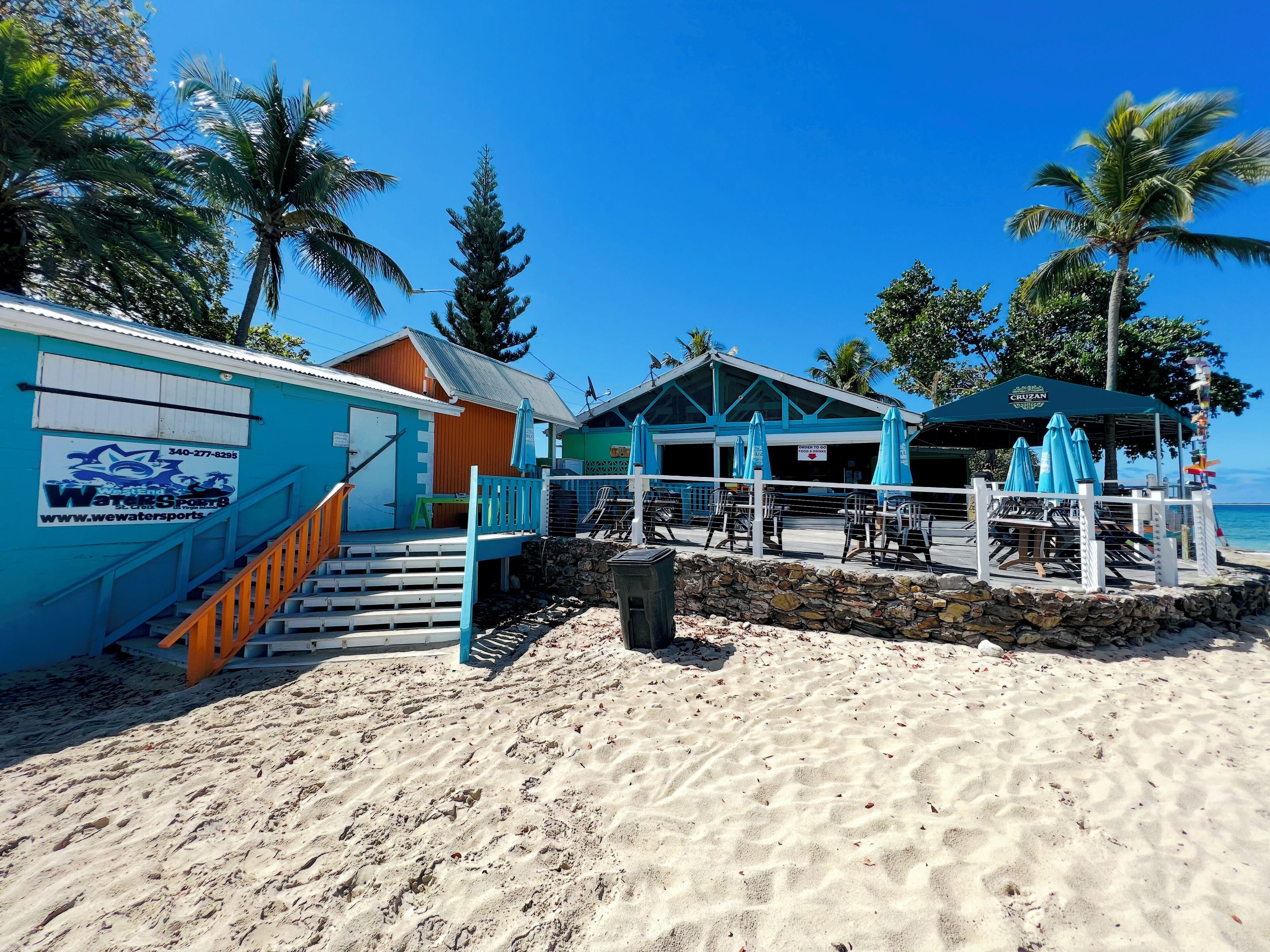 A beachside building with a staircase leading to a deck area, featuring outdoor seating and a view of the ocean.