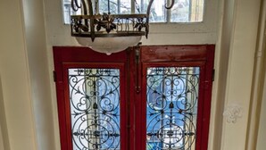Eine rote Tür mit aufwendiger Metallverzierung und Glasschilden, ein gemustertes Parkett und ein kleiner Hocker auf der rechten Seite.