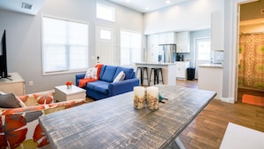 Una sala de estar moderna con un sofá azul, una mesa de centro de madera y una zona de cocina con armarios blancos y un refrigerador de acero inoxidable.