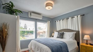 Ein Schlafzimmer mit Bett, Nachttisch, Lampe, einem Fenster mit Vorhängen und einer Klimaanlage.