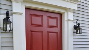 A red door with a black doormat and two wall-mounted lanterns.