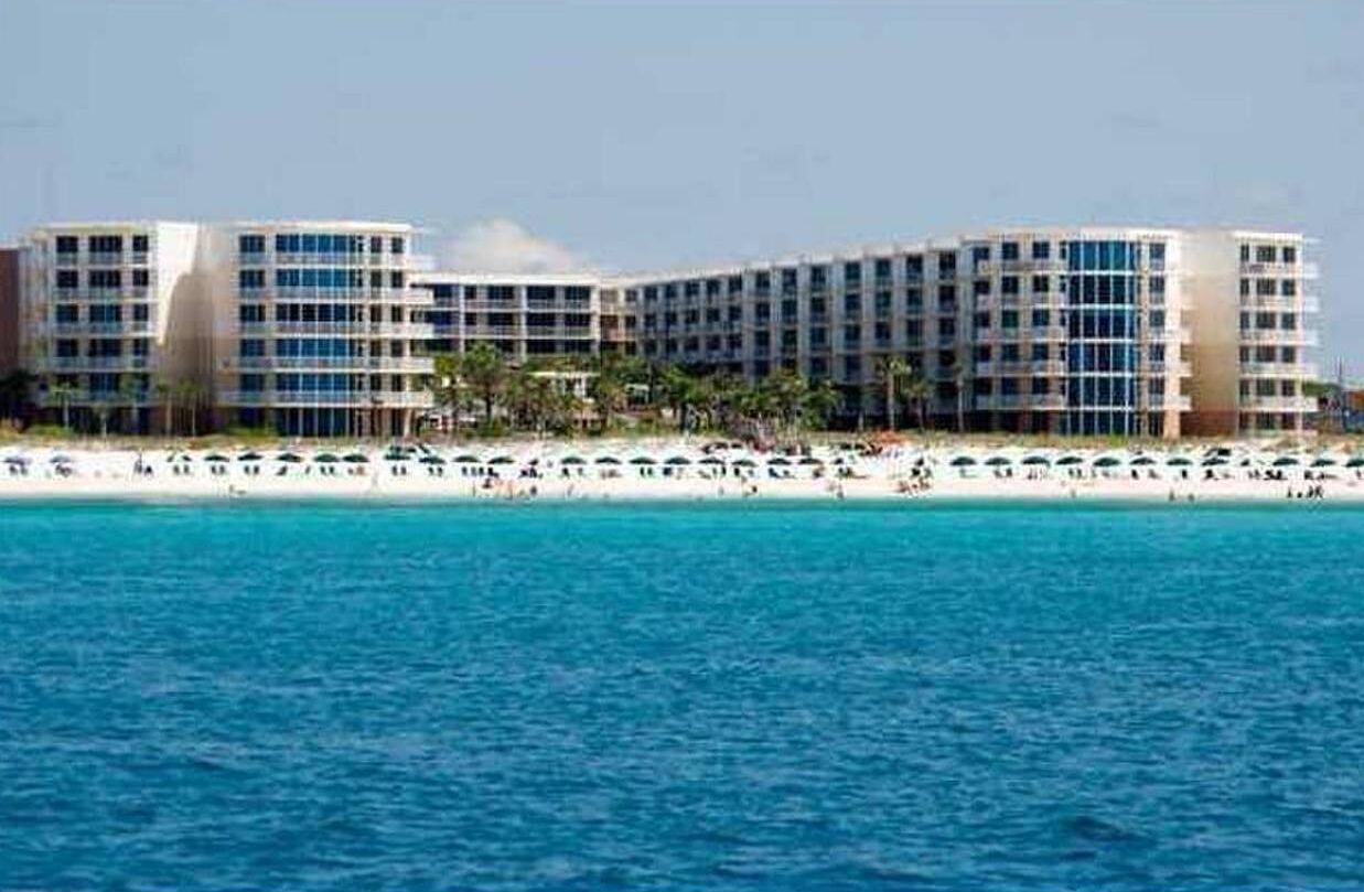 Un grande hotel con diversi piani e balconi, situato vicino a una spiaggia con sabbia fine e acqua cristallina.