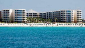 A large hotel with multiple floors and balconies, located near a beach with a sandy shore and clear blue water.