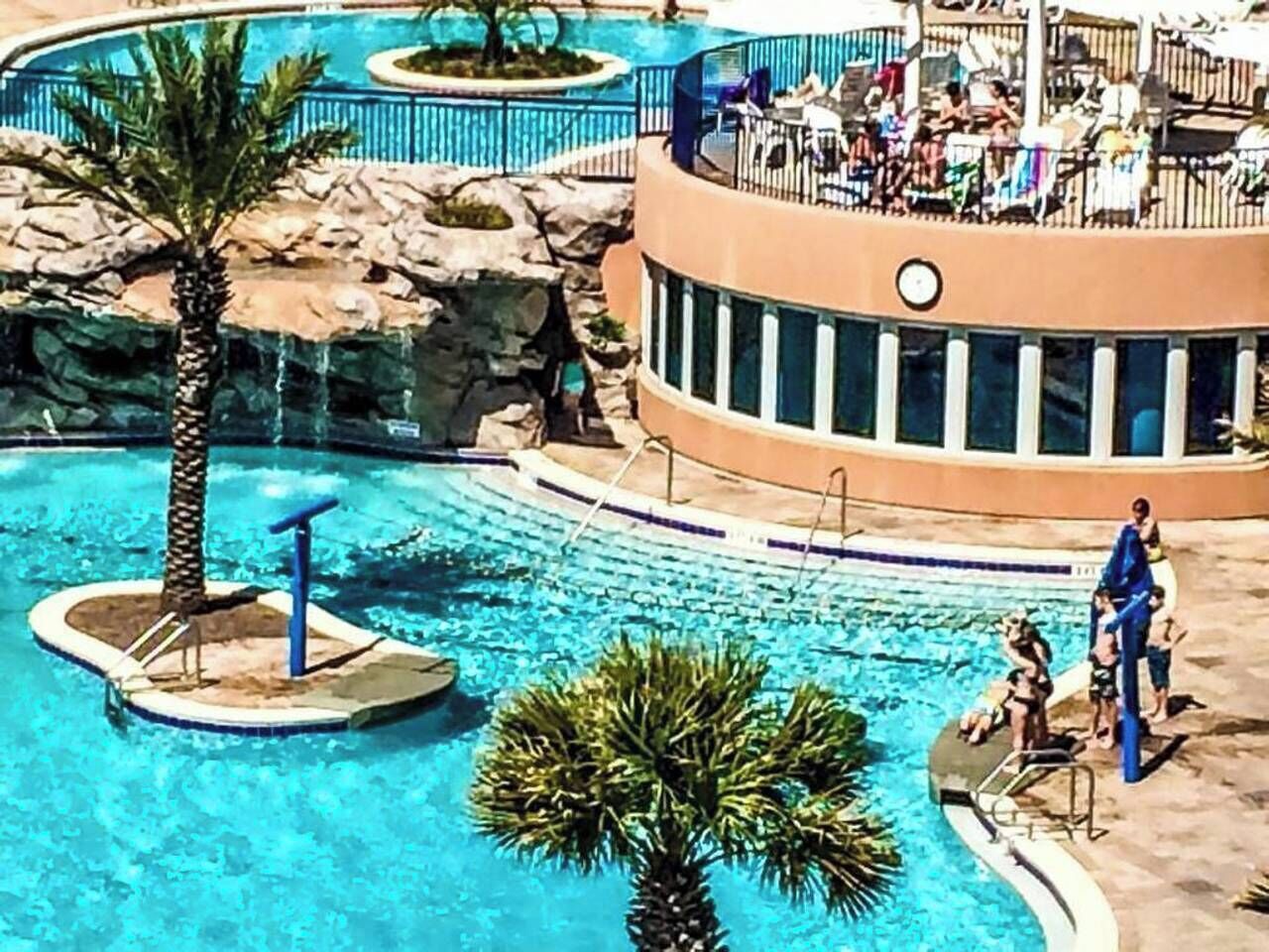 Ett poolområde med en palm, en liten ö med en vattenfontän och en loungehörna med folk.
