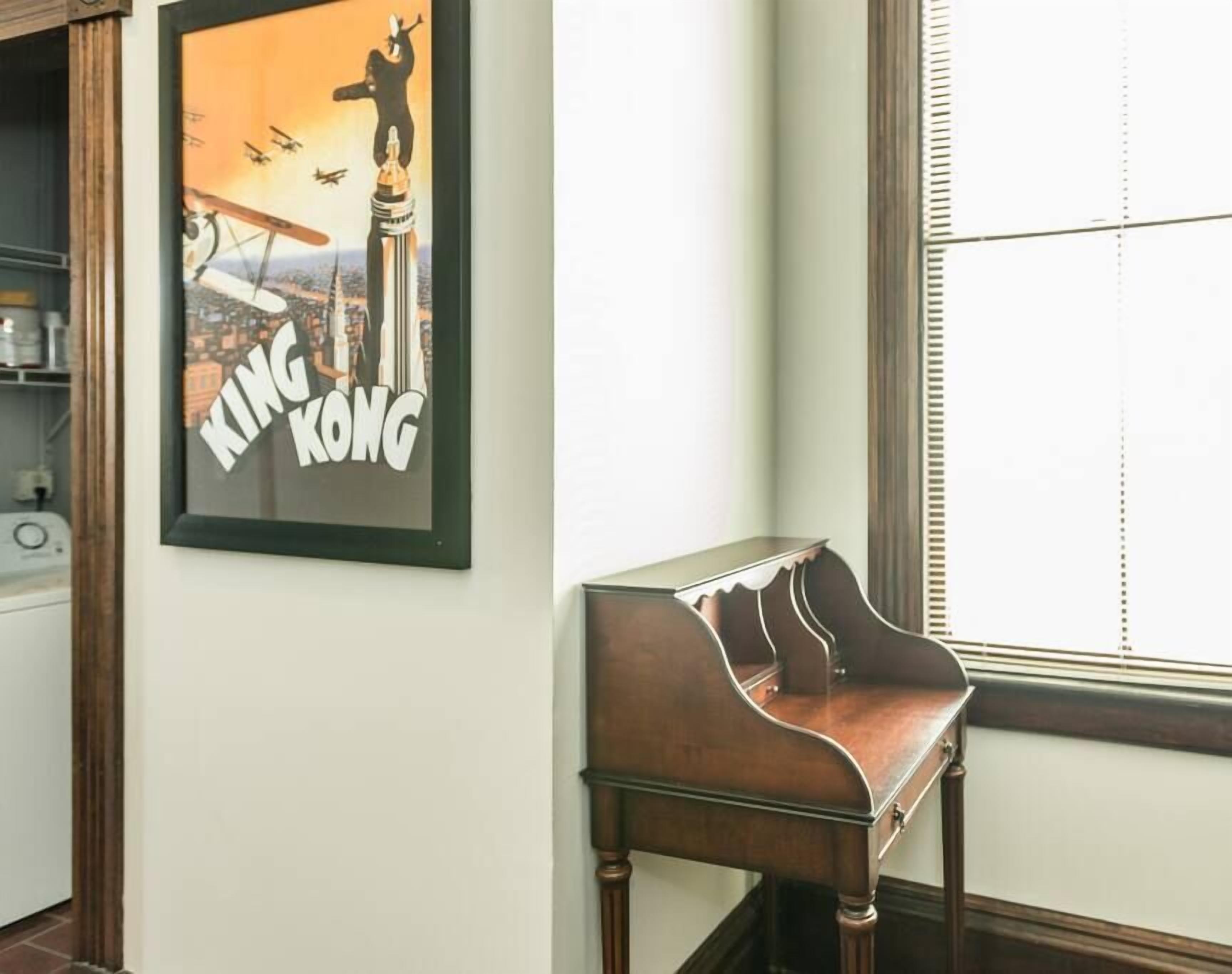 A framed poster of King Kong on a wall, a wooden piano, and a window with blinds.