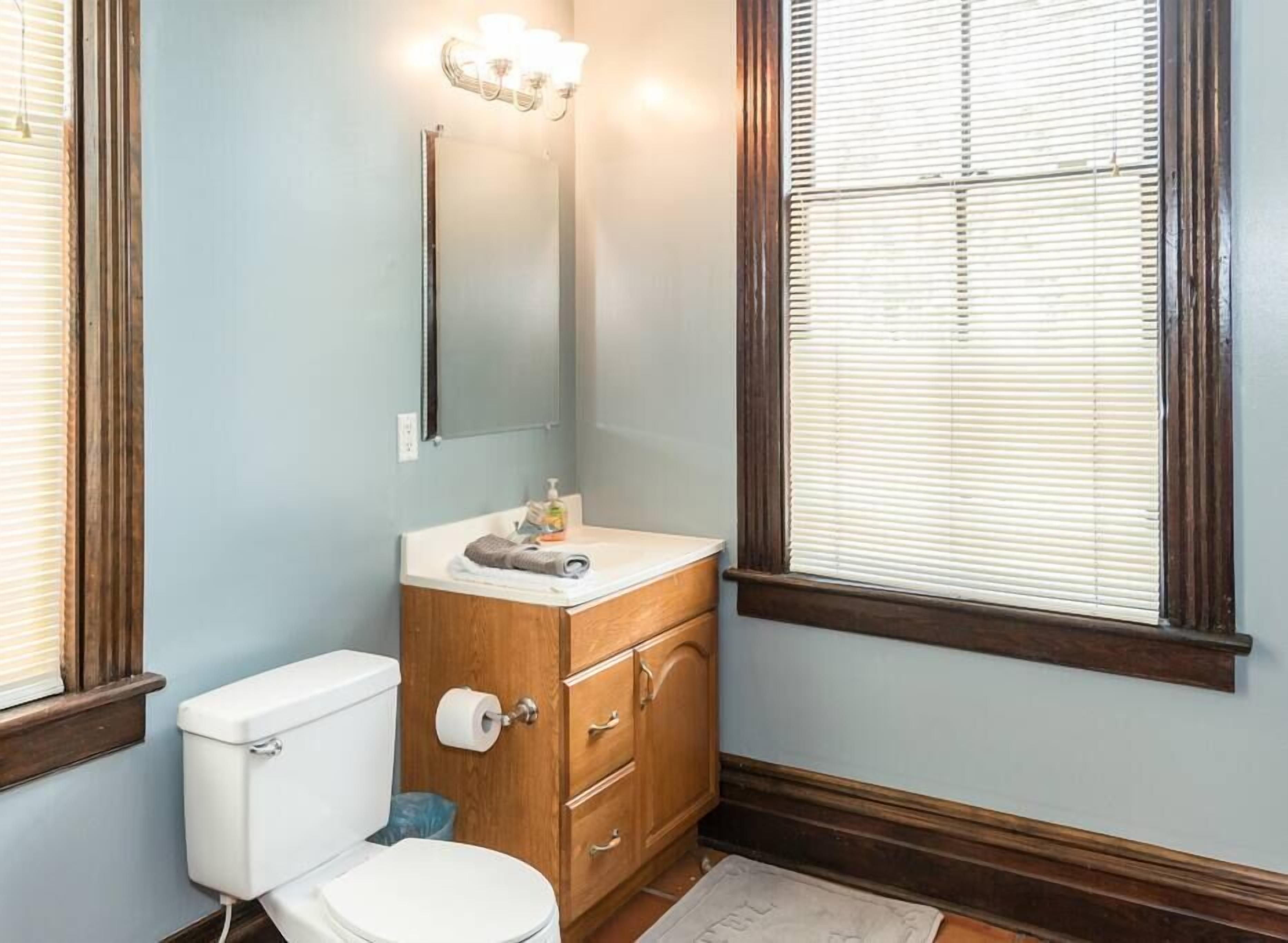Ein Badezimmer mit einem Holzkosmetikschrank, einer Toilette, einem Spiegel und einem Fenster mit Jalousien.