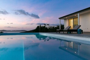 A modern infinity pool with clear water reflecting the sky, two lounge chairs, and a building with large windows.