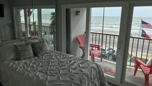 Una camera da letto con un letto grande, un balcone con sedie rosse e vista sulla spiaggia e sull'oceano.