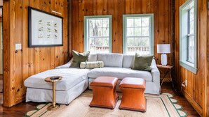 A wooden-paneled room with a sectional sofa, two ottomans, a framed picture, and two windows. - Riverfront House With Dock Sleeps 8 (St Simons Island)