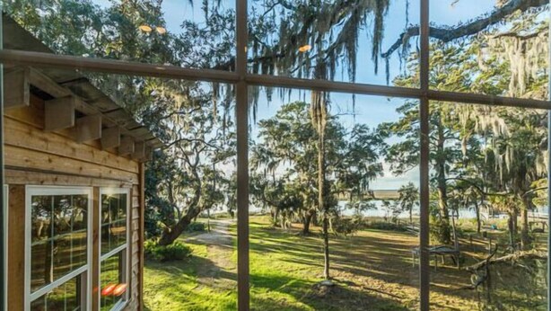 A wooden cabin with large windows overlooking a grassy yard with trees and a body of water. - Riverfront House With Dock Sleeps 8 (St Simons Island)