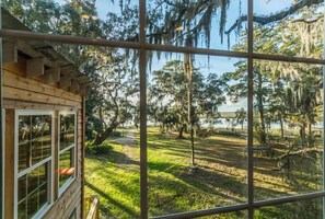 A wooden cabin with large windows overlooking a grassy yard with trees and a body of water. - Riverfront House With Dock Sleeps 8 (St Simons Island)