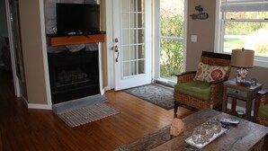Una sala de estar con chimenea, televisor, una silla de mimbre, una mesa de centro y un ventanal con persianas.