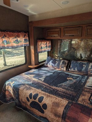A bedroom with a large bed, a window with curtains, and a wall-mounted TV. - Sandpiper 40ft RV @EvansCliff /HotTub on Covered Deck! new 2026 location (Hot Springs)