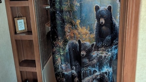A bathroom with a shower curtain depicting bears in a forest, wooden shelves, and a small bedside table. - Sandpiper 40ft RV @EvansCliff /HotTub on Covered Deck! new 2026 location (Hot Springs)