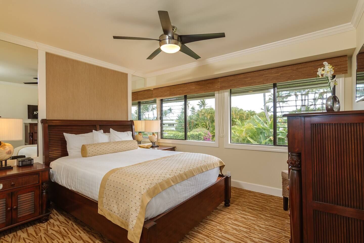 A spacious bedroom with a large bed, wooden headboard, a ceiling fan, and a view of lush greenery through the windows.