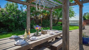 A wooden pergola with a table set for dining, surrounded by a garden with flowers and a fence in the background.