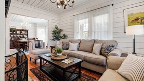 A spacious living room with a large sofa, a coffee table, and a chandelier. There is a dining area with a table and chairs visible in the background.