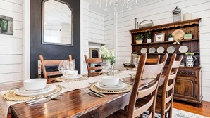 A dining room with a wooden table set for six, surrounded by chairs. A chandelier hangs from the ceiling, and a wooden cabinet with decorative plates and a birdcage is visible.