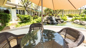 A glass-top table with wicker chairs, a beige umbrella, and a small waterfall in a landscaped garden.
