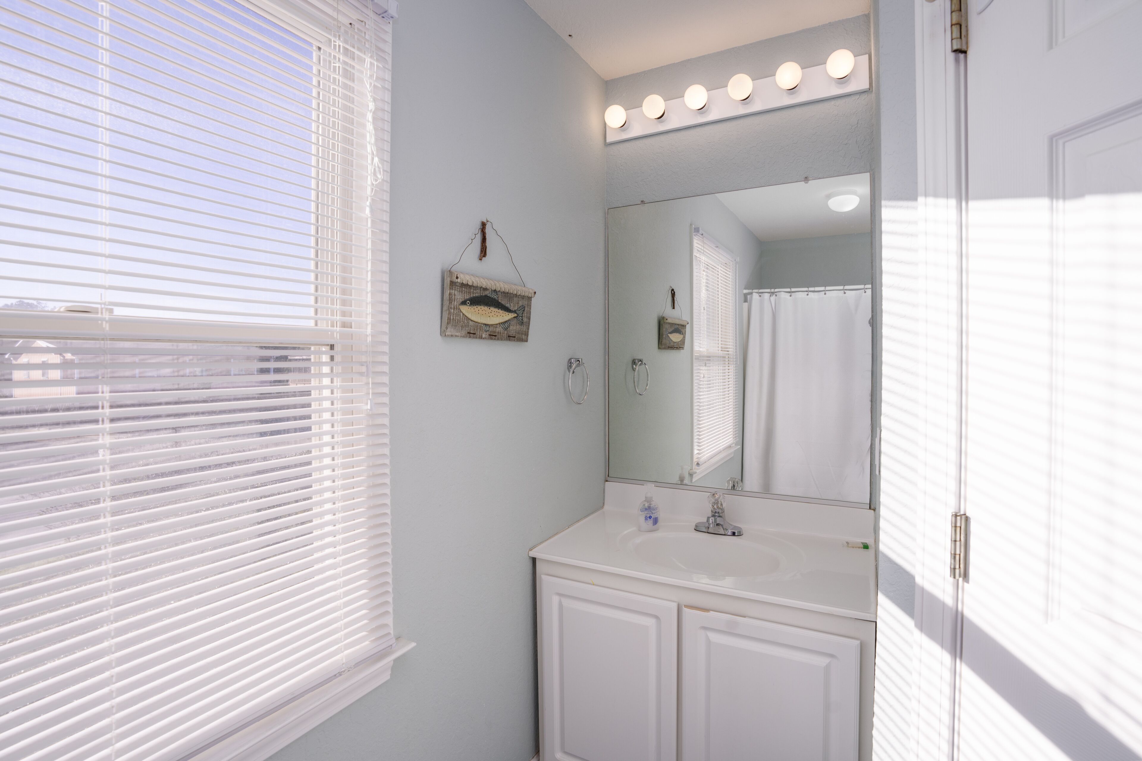 A bathroom with a white vanity, a large mirror with lights, and a window with blinds.