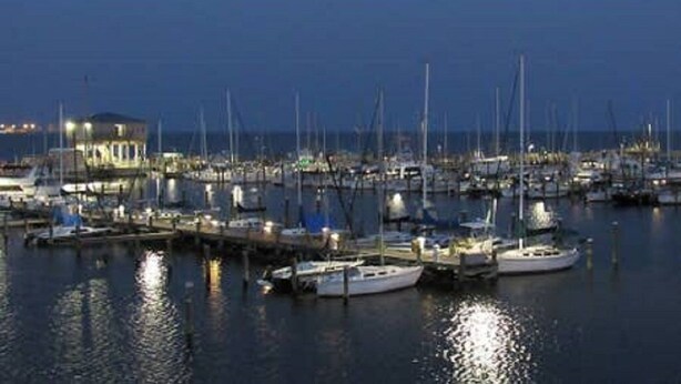 A marina at dusk with multiple boats docked, a building with lights, and calm water reflecting the lights.