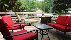 A wooden deck with red cushioned chairs and a small table, overlooking a dock and a house.