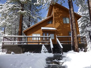 Una cabaña de madera con un balcón y una escalera que lleva a la entrada, rodeada de árboles cubiertos de nieve.