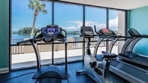 Ett gym med tre löpband och en motionscykel, stora fönster med utsikt över en palm och vattnet.