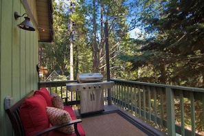 A covered outdoor deck with a grill, red cushioned seating, and a view of a forest.