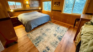 A cozy wooden cabin bedroom with a bed, a rug, and a window with a view of snow-covered trees.