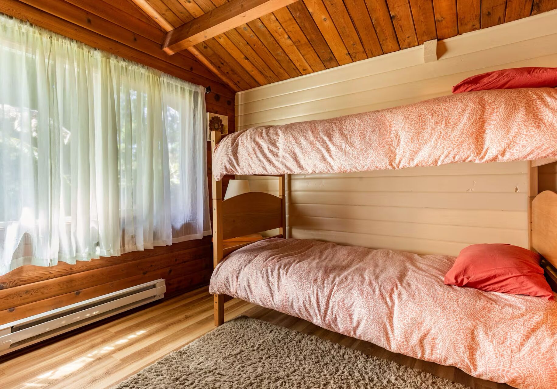 A bunk bed room with wooden walls, a window with sheer curtains, and a radiator.