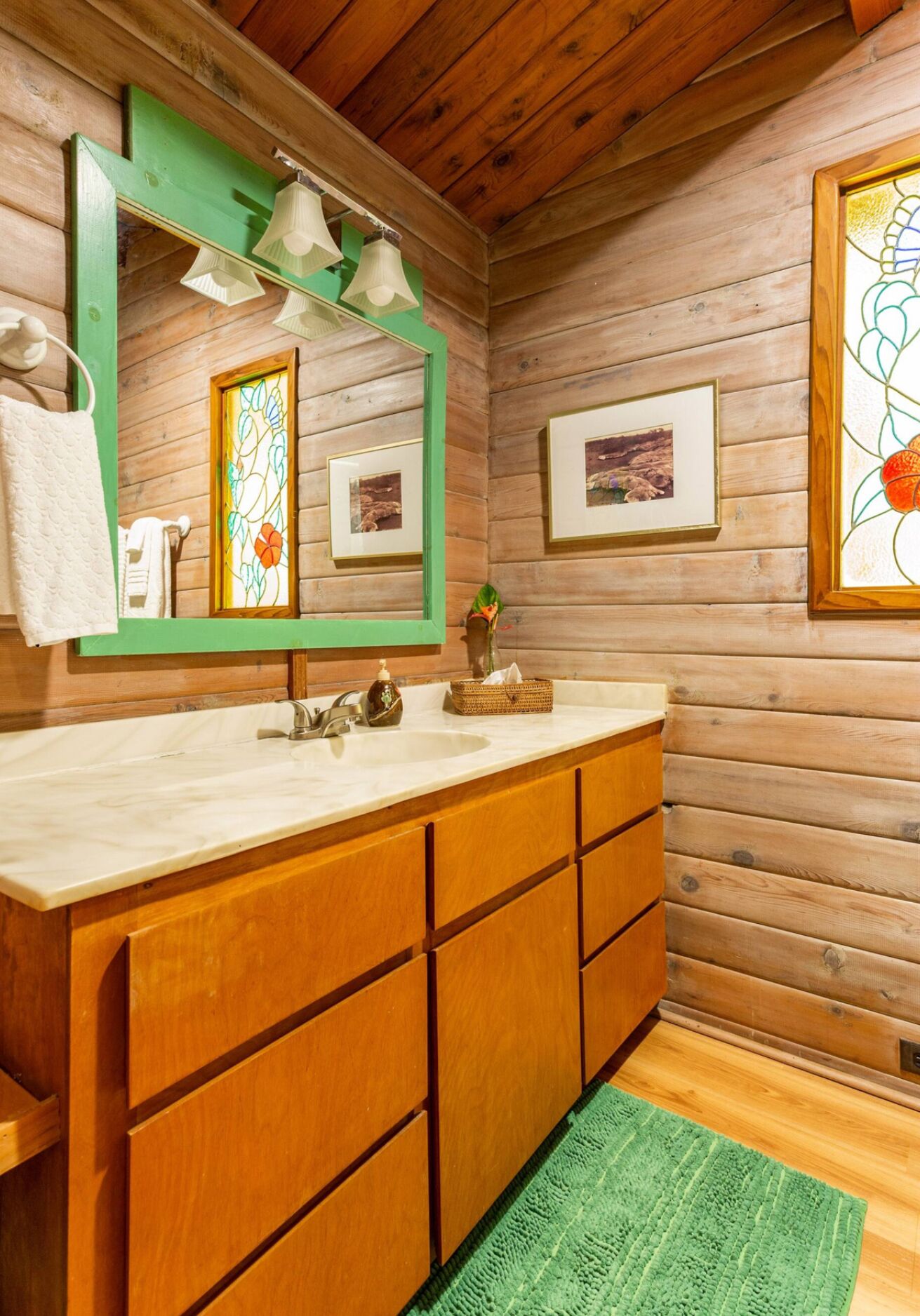 A wooden cabin bathroom with a large wooden vanity, a green mirror, and framed pictures on the walls.
